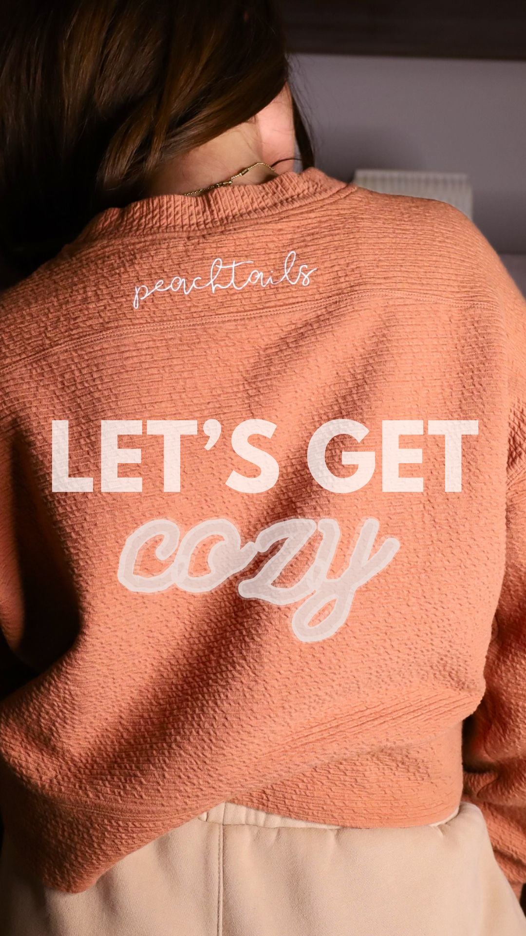 The image captures the back of a woman wearing a cozy, pink sweater with the words "LET'S GET cozy" in large, white cursive text overlaying the photo. The sweater's texture is visible, suggesting softness and warmth. In the top left corner, the word "peachtails" is scripted in a smaller, delicate font. The lighting and composition of the photo convey a comfortable and snug atmosphere, inviting the viewer to embrace the concept of getting cozy.