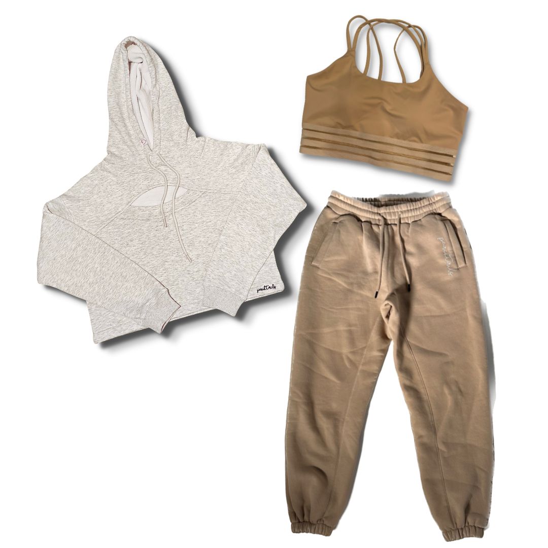 The image displays a set of athleisure wear: a heather grey crop hoodie with a hood, a tan sports bra with multiple straps, and matching light mocha joggers. The pieces are laid out separately against a white background to showcase their design and color.