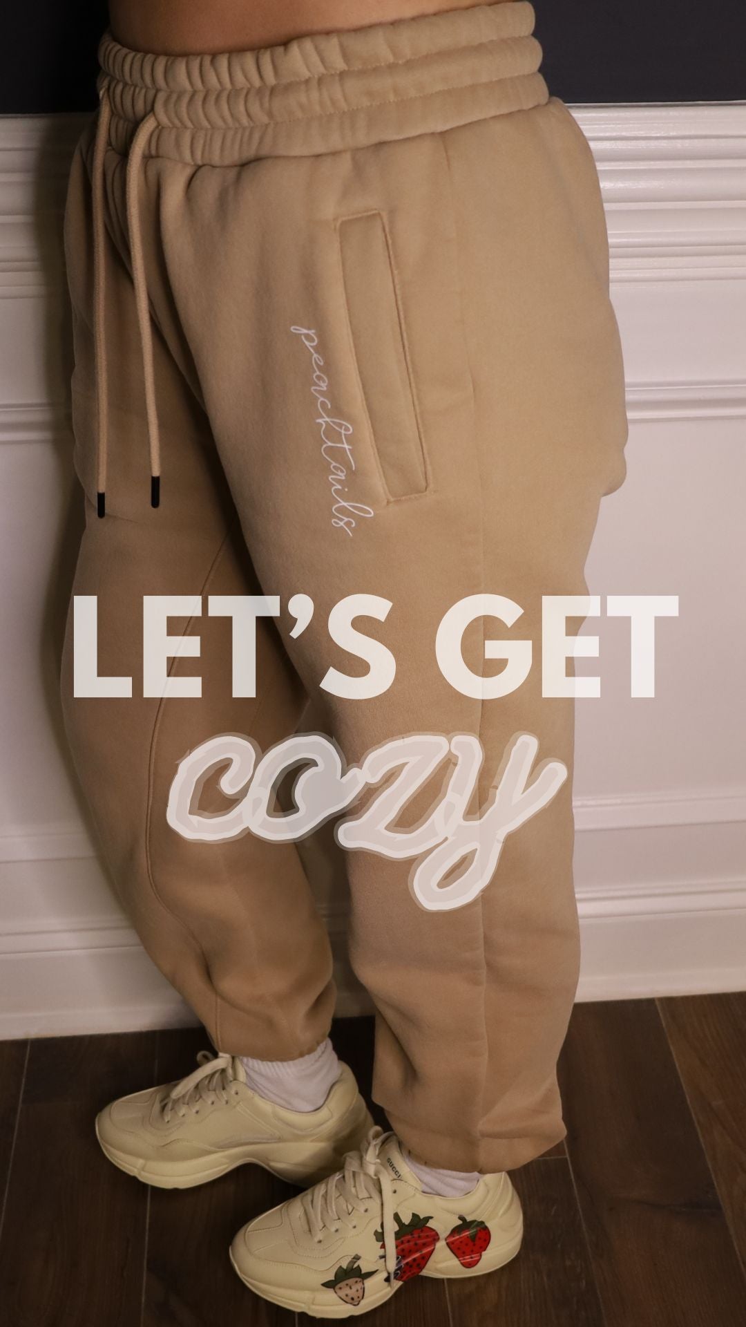 The image shows a close-up of someone wearing beige, cozy joggers with the word "peachtails" embroidered on them. Below, the phrase "LET'S GET cozy" in white cursive lettering overlays the image. The person is also wearing cream-colored sneakers with a red heart design. The focus on the soft joggers and comfortable footwear emphasizes a relaxed and comfortable style, inviting the viewer to enjoy a cozy experience.