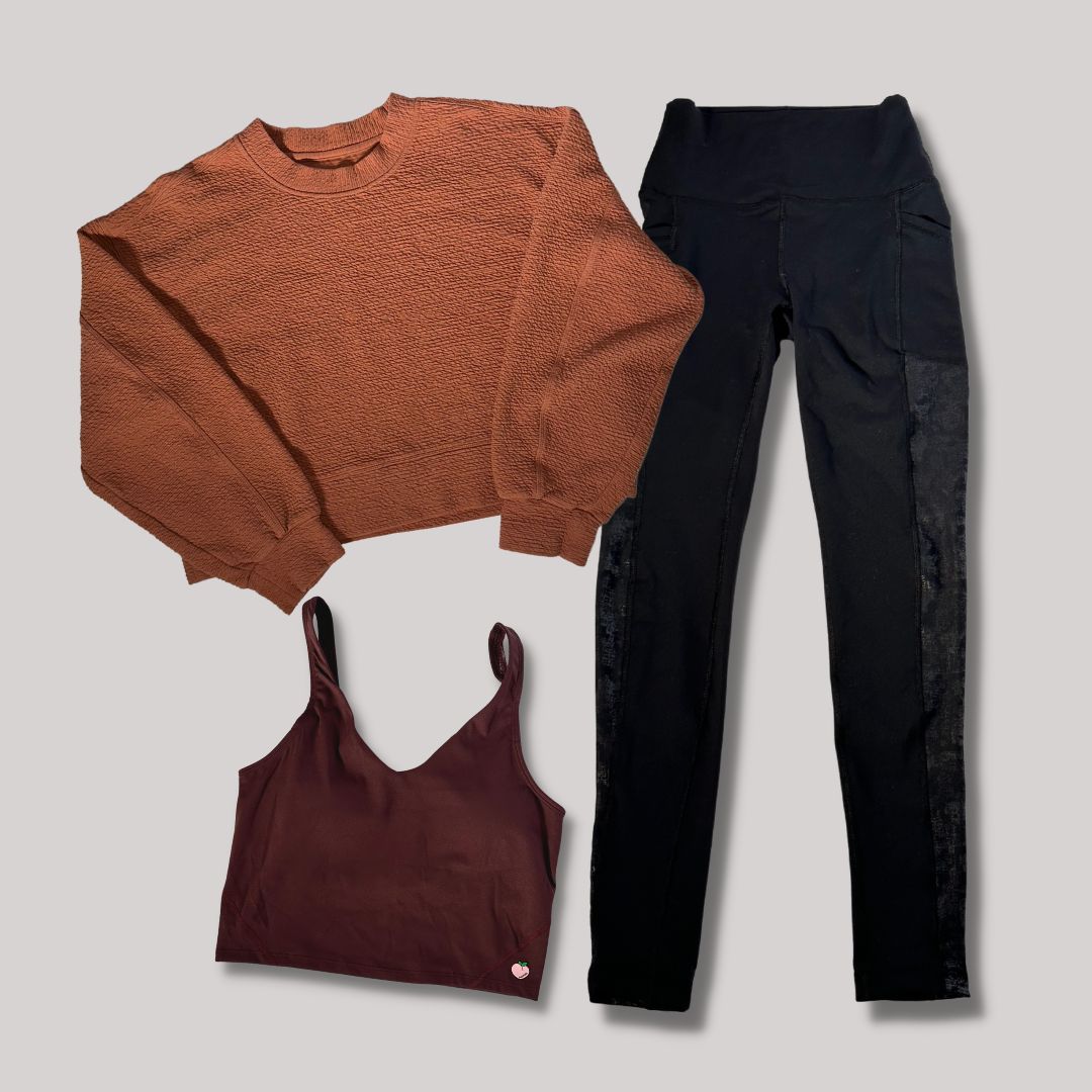 The image shows a collection of women's clothing laid out on a white background: a cinnamon-colored crewneck sweater with puffed sleeves, a dark burgundy sports bra, and black joggers with lace detailing along the sides. The arrangement highlights the garments' colors and styles.