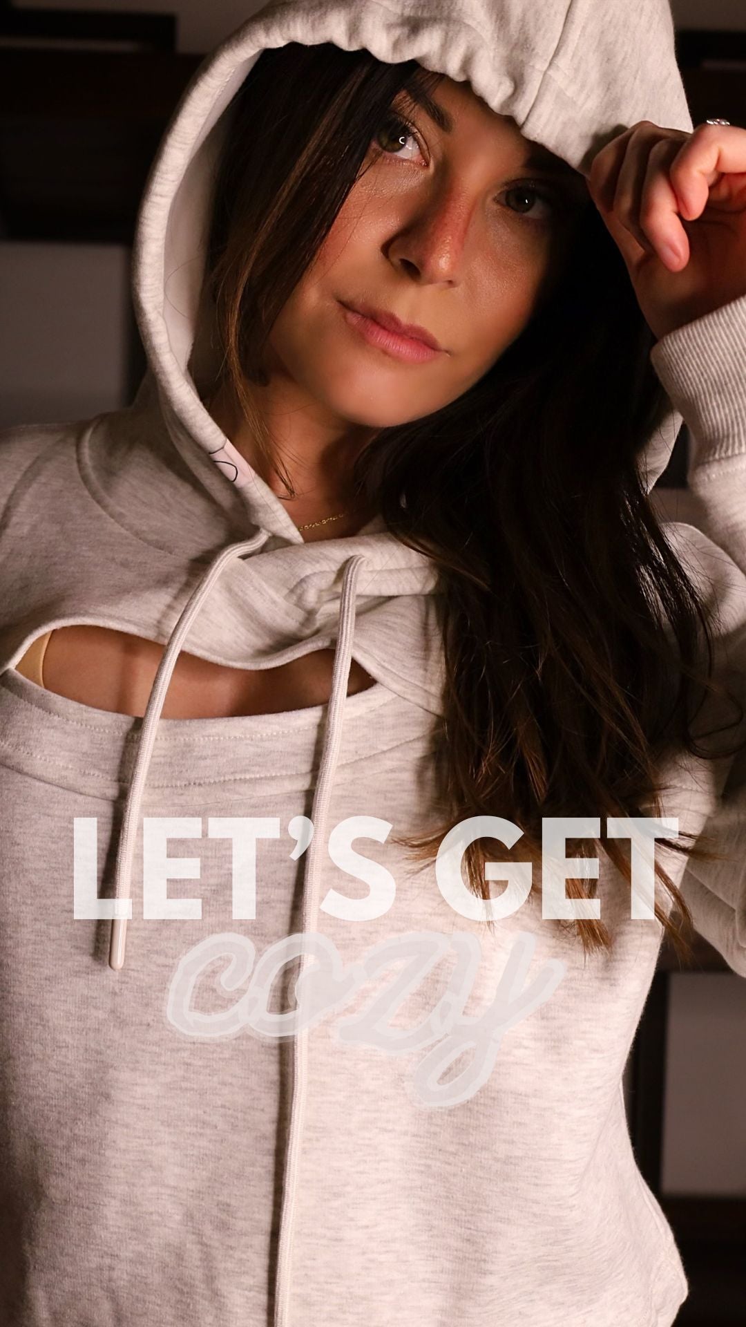 The image features a woman pulling the hood of a light grey hoodie over her head, with the phrase "LET'S GET cozy" in white cursive lettering across the image. Her gaze is directed towards the camera, with a relaxed and comfortable expression that matches the cozy theme suggested by the hoodie and the overlaid text. The intimate and warm atmosphere of the photo invites viewers to embrace comfort and relaxation.