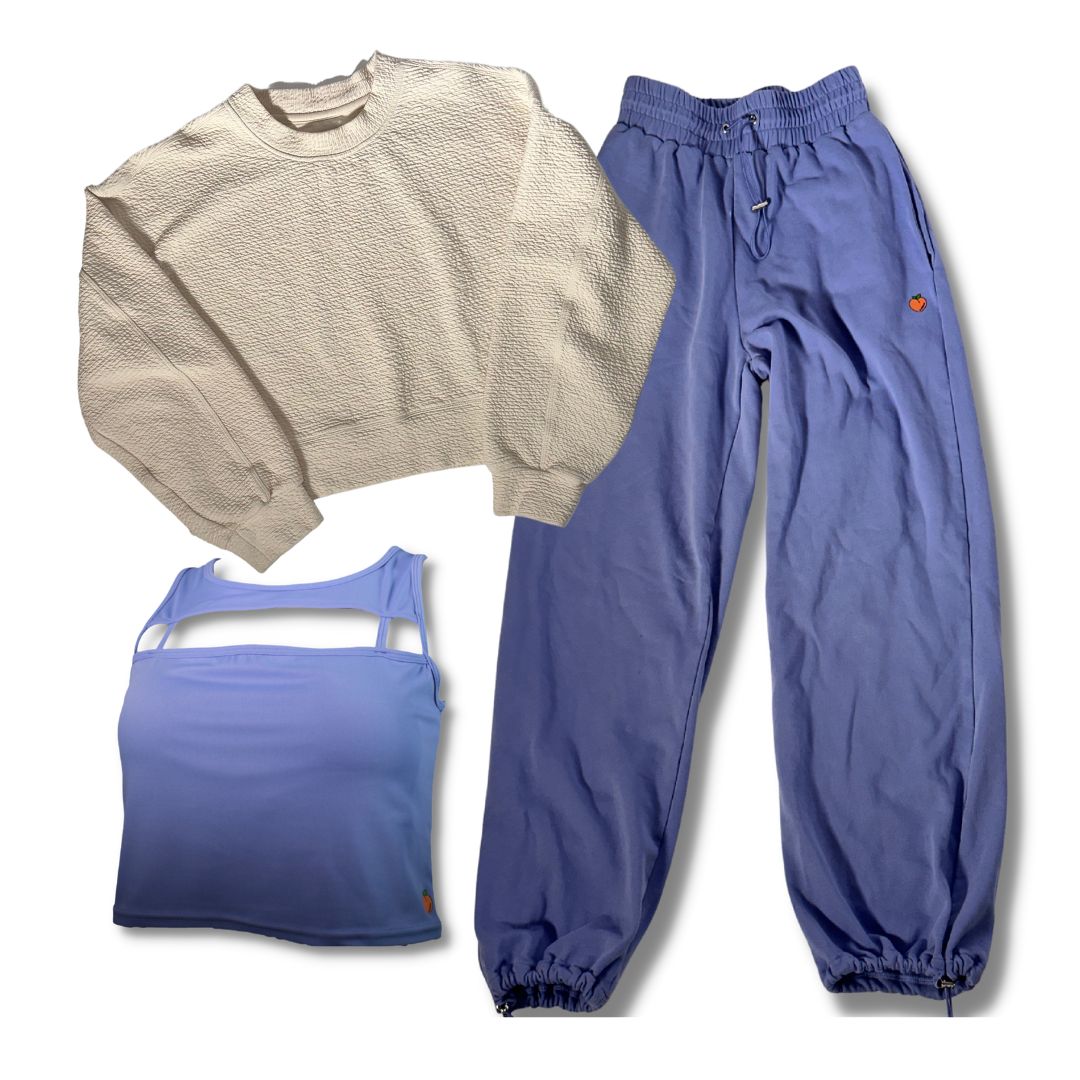 The image shows a set of women's athleisure clothing laid out against a white background, featuring a vanilla bean-colored crewneck sweater, a light blue sports tank top, and periwinkle blue joggers with elastic cuffs. Each piece is placed to showcase its design and fit.