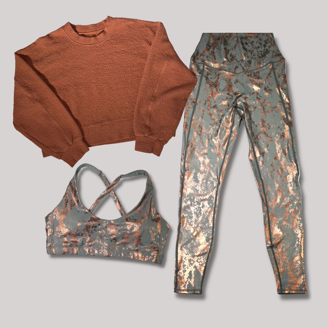 The image features a cinnamon-colored crewneck sweater, a sports bra with a crisscross back in a matching camo-like pattern, and leggings with the same design. The leggings and bra have metallic accents, and the clothing is displayed against a white background.